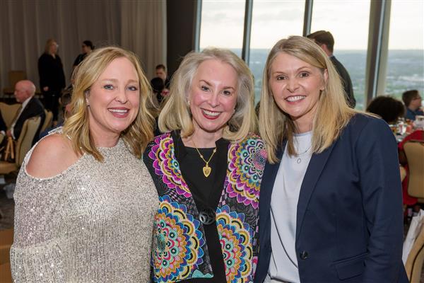 Three women wearing business casual attire smile at the camera. They all have blond hair and a pleasant smile. 